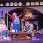 Dame Maureen Lipman as Allegra stands with her arms raised while Elizabeth Bower as Anna and John Middleton as Ronen sit in the cottage.