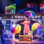 Dame Maureen Lipman as Allegra stands beneath rainbow umbrellas while Bailey Patrick as Officer Rogers watches during a colourful musical sequence.
