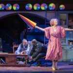 Dame Maureen Lipman as Allegra performs with a closed rainbow umbrella while John Middleton as Ronen and Bailey Patrick as Officer Rogers sit behind her.