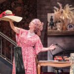 Dame Maureen Lipman as Allegra holding up a wide-brimmed hat in the cottage sitting room.